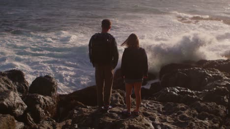 A-couple-stands-on-a-rocky-shore-as-waves-roll-in-and-crash