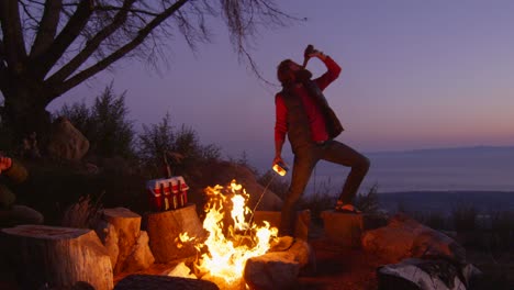 Ein-Mann-Sprüht-Feuerzeugflüssigkeit-Auf-Ein-Lagerfeuer,-Während-Er-Auf-Einem-Campingplatz-Bier-Trinkt