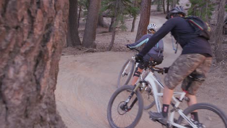 The-camera-pans-around-a-tree-trunk-to-reveal-two-mountain-bikers-riding-on-a-dirt-path-in-a-forest