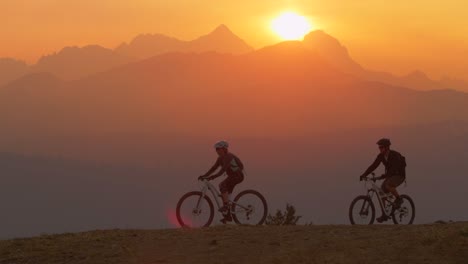 Two-mountain-bikers-pull-to-a-stop-on-a-hillside-at-sunset
