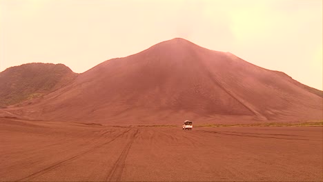 Ein-Fahrzeug-Fährt-Durch-Eine-Wüstenlandschaft,-Während-Sich-Im-Hintergrund-Eine-Vulkanaschewolke-Bildet