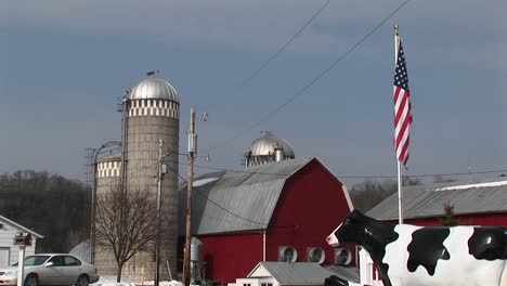 The-Camera-Captures-An-American-Farm-With-Silos-Red-Barn-American-Flag-And-Lifesize-Replica-Of-A-Cow
