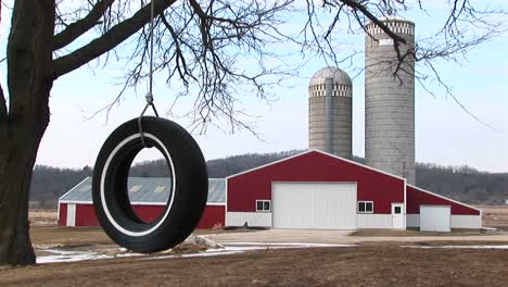 A-Tire-Swing-Hangs-From-A-Tree-With-Farm-Buildings-And-Twin-Silos-In-The-Background