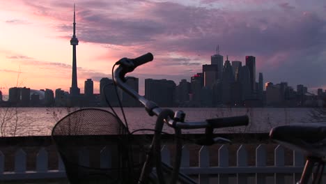 A-Bicycle-Is-Parked-Near-The-Coastline-Of-Lake-Ontario-With-The-Skyline-Of-Toronto-In-The-Background