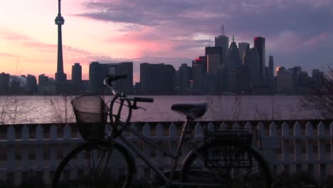Un-Turista-Quita-Una-Bicicleta-Estacionada-En-Una-De-Las-Islas-De-Toronto-En-Goldenhour-Con-El-Lago-Ontario,-La-Torre-Y-El-Horizonte-De-Toronto-En-La-Distancia