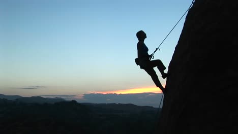 Longshot-Of-A-Rock-Climber-Silhouetted-Against-A-Goldenhoursky-Rappelling-Down-A-Cliff