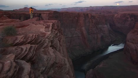 Weitwinkelaufnahme-Eines-Fotografen,-Der-Auf-Einer-Klippe-Steht-Und-Die-Canyons-Des-Colorado-River-Fotografiert,-Während-Er-Sich-Unter-Einem-Aufgehenden-Vollmond-Durch-Enge-Sandsteinschluchten-Schlängelt