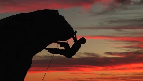 Mediumshot-Of-A-Silhouetted-Climber-Hanging-From-An-Overhang-Rock-Face-With-A-Beautiful-California-Sunset-In-The-Distance