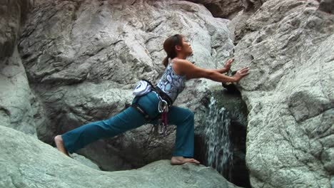 Mediumshot-Of-A-Woman-Doing-Stretches-Next-To-A-Small-Waterfall-On-A-Granite-Rockface