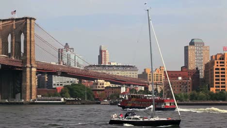 Eine-Fähre-Fährt-Unter-Der-Brooklyn-Bridge-In-New-York-City-Durch,-Während-Ein-Segelboot-Vorbeifährt
